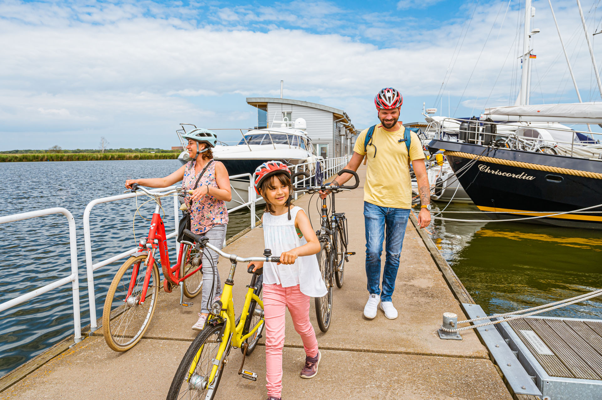 Kind schiebt gelbes Fahrrad am HAfen entlang. ImHintergrund die Ektern mit Fahrrädern. links und rechts ist Wasser und Boote, die fest gemacht sind.